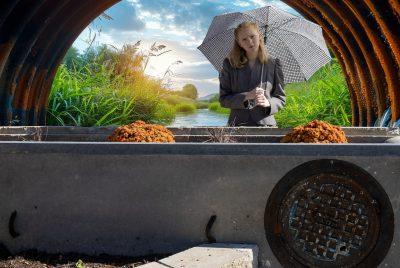 A woman holding an umbrella standing in front of a tranquil stream gently flowing through a metal culvert amidst lush greenery